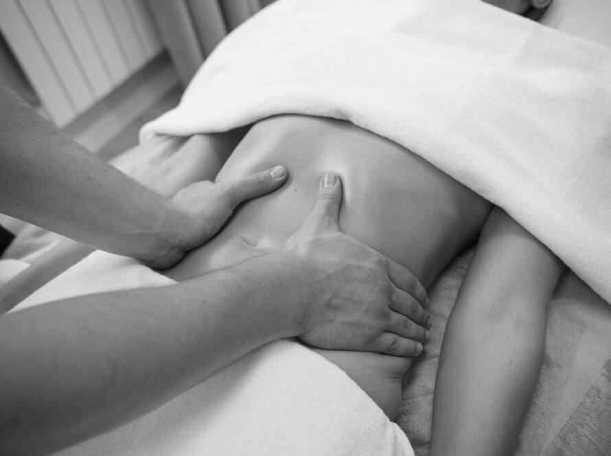 Black and white image of a client face down and the massage therapist has his hands around the upper waist of the client whilst using his thumbs to make deep strokes up the middle of the back during a Lymphatic drainage massage.