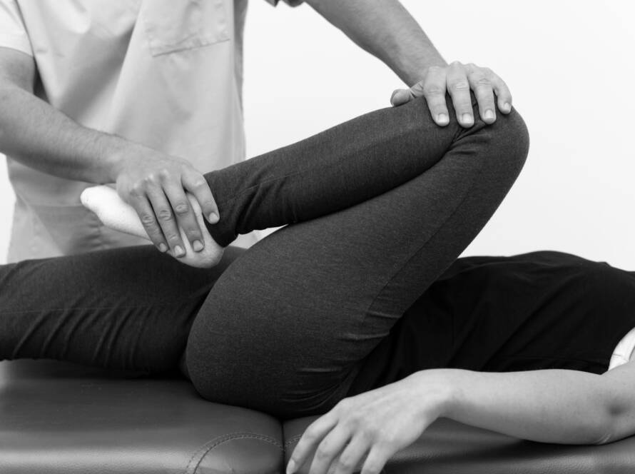 black and white image of a client on her back with her left knee brought up towards her chest, whilst the therapist applies pressure to the knee and the ankle as part of a sports rehabilitation program.