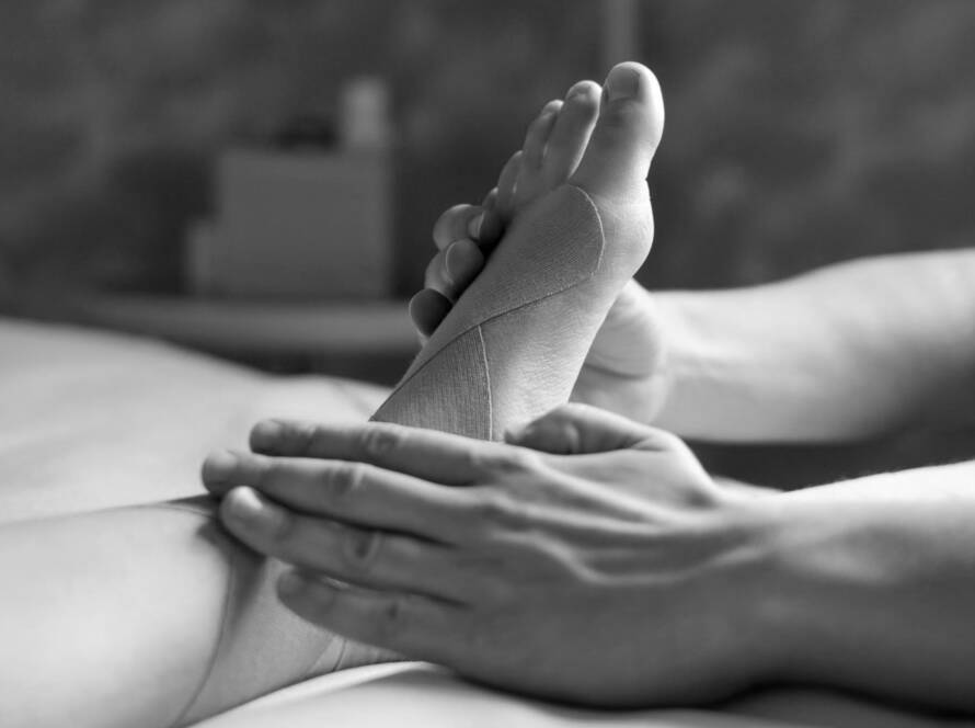Black and white image of a foot with Kinesio tape on it. The massage therapist has both hands on the foot and is using his thumb to massage the sole of the foot, during a sports therapy massage.